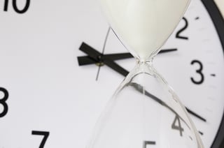 Two measurements of time White sand falling inside hourglass, with round analog clock in  background (focus on neck of hourglass), shallow depth of field.jpeg